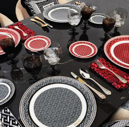 Versace luxury dinnerware in black and red Greek key pattern with gold cutlery and black crystal glasses on a marble table.