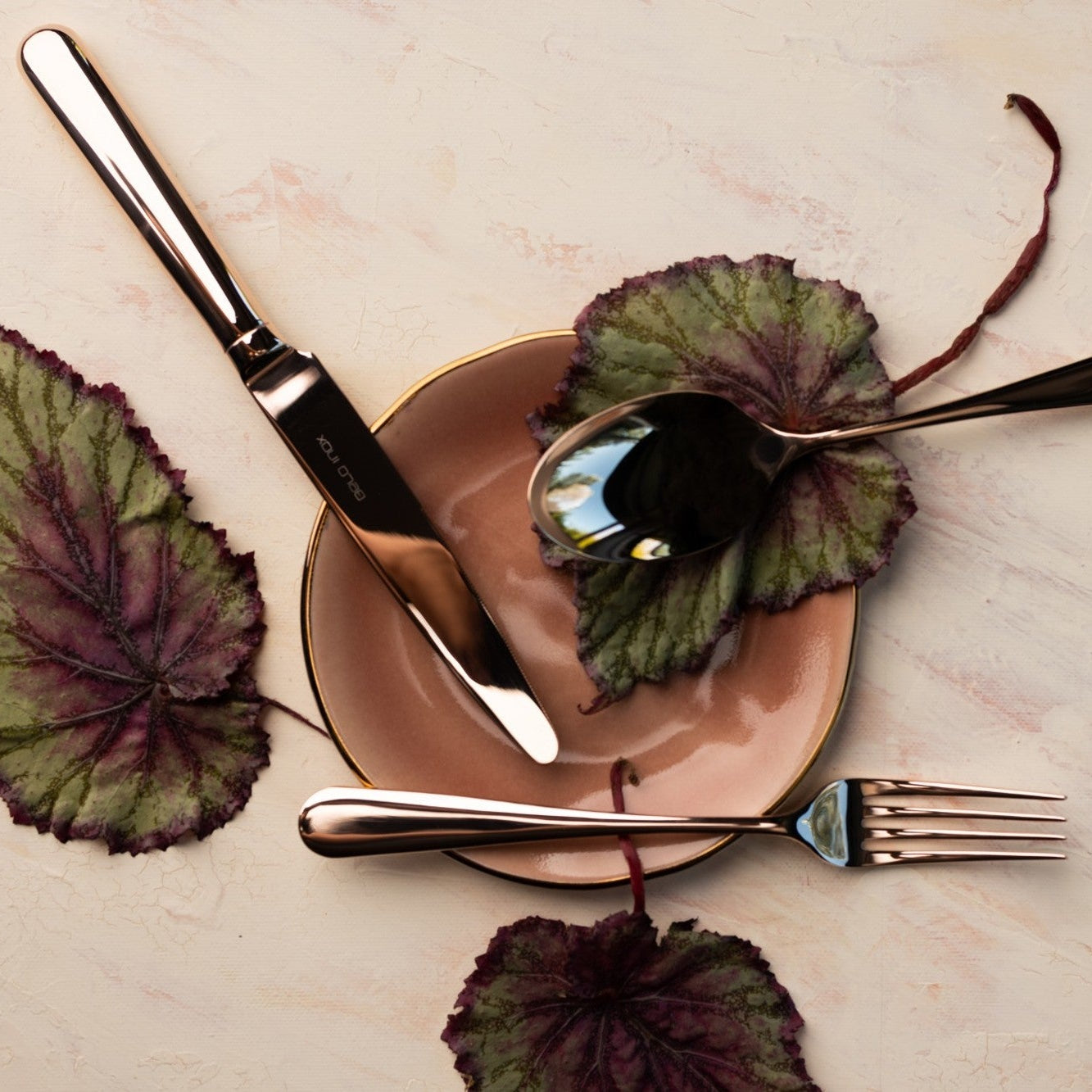 Belo Inox Belo 9 rose gold cutlery set displayed on a blush plate with decorative leaves – elegant luxury table setting