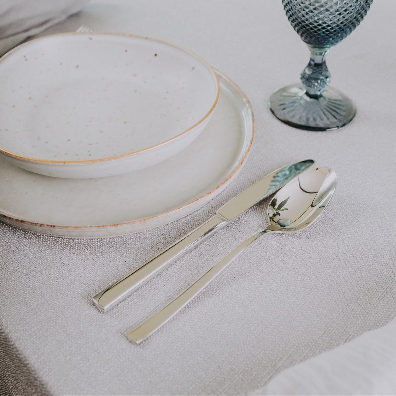 Belo Inox Belo 12 cutlery set elegantly arranged on a neutral table setting with ceramic plates and blue glass