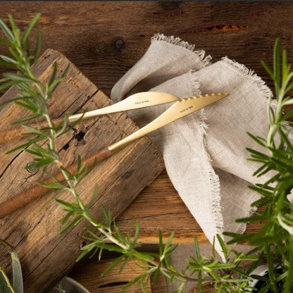 Gold table knife and steak knife from the Belo Inox Neo Brown collection on a rustic wooden board with fresh herbs and a linen napkin