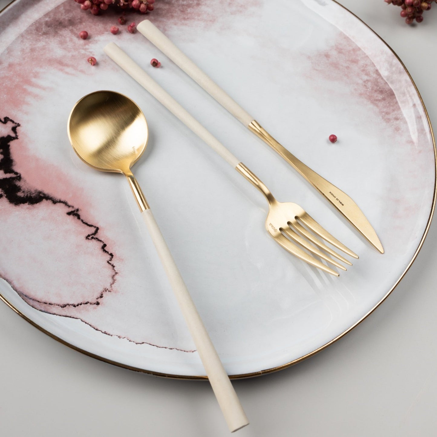 Belo Inox NEO Gold cutlery set placed on a pink and white ceramic plate with decorative details, lifestyle table setting.
