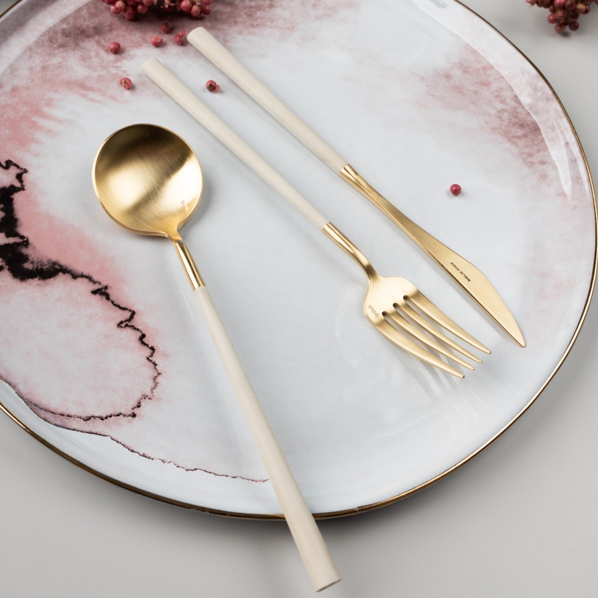 Belo Inox NEO Gold cutlery set placed on a pink and white ceramic plate with decorative details, lifestyle table setting.
