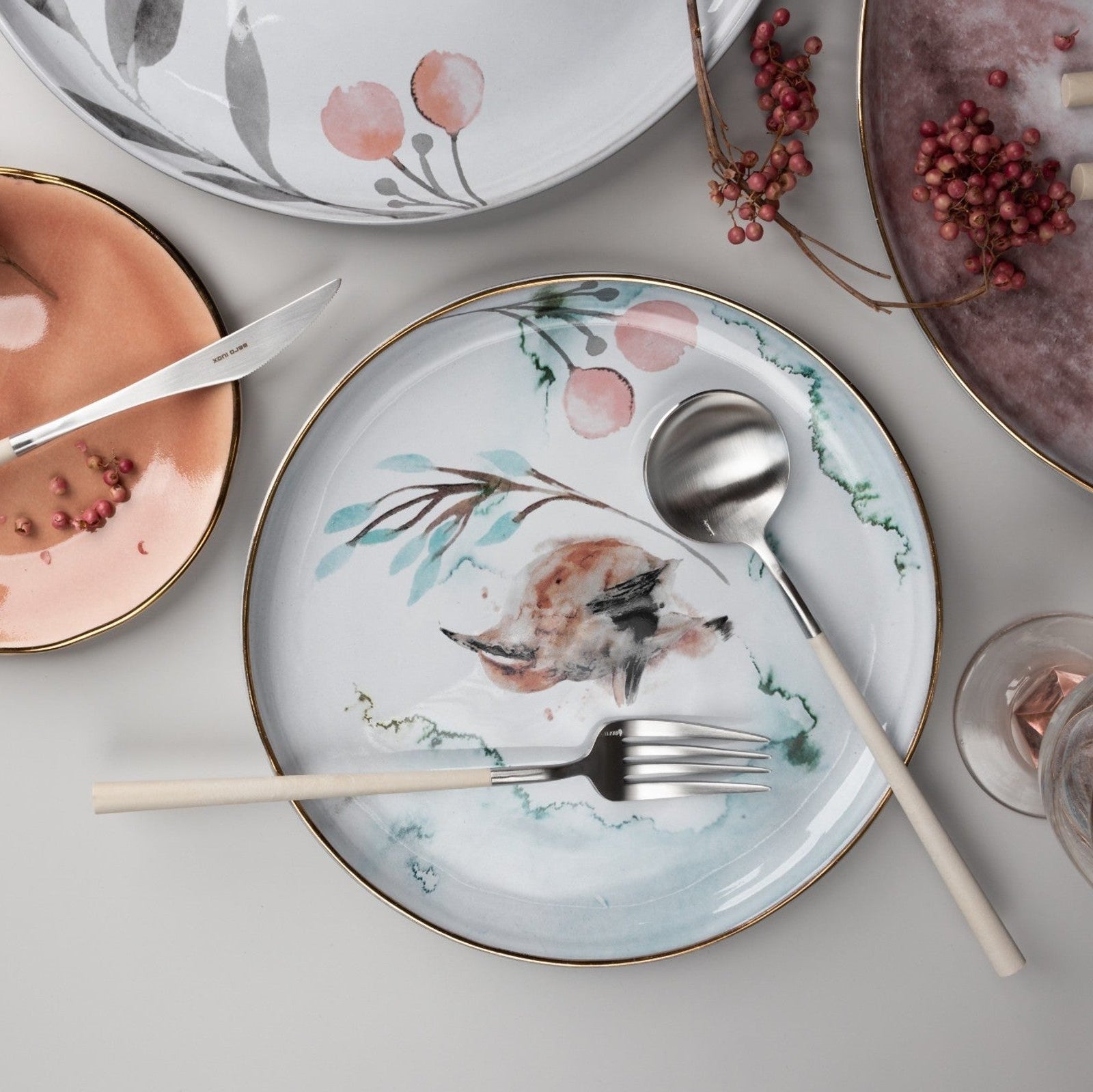 Belo Inox NEO Ivory cutlery set displayed on floral ceramic plates with a light grey background, lifestyle table setting.

