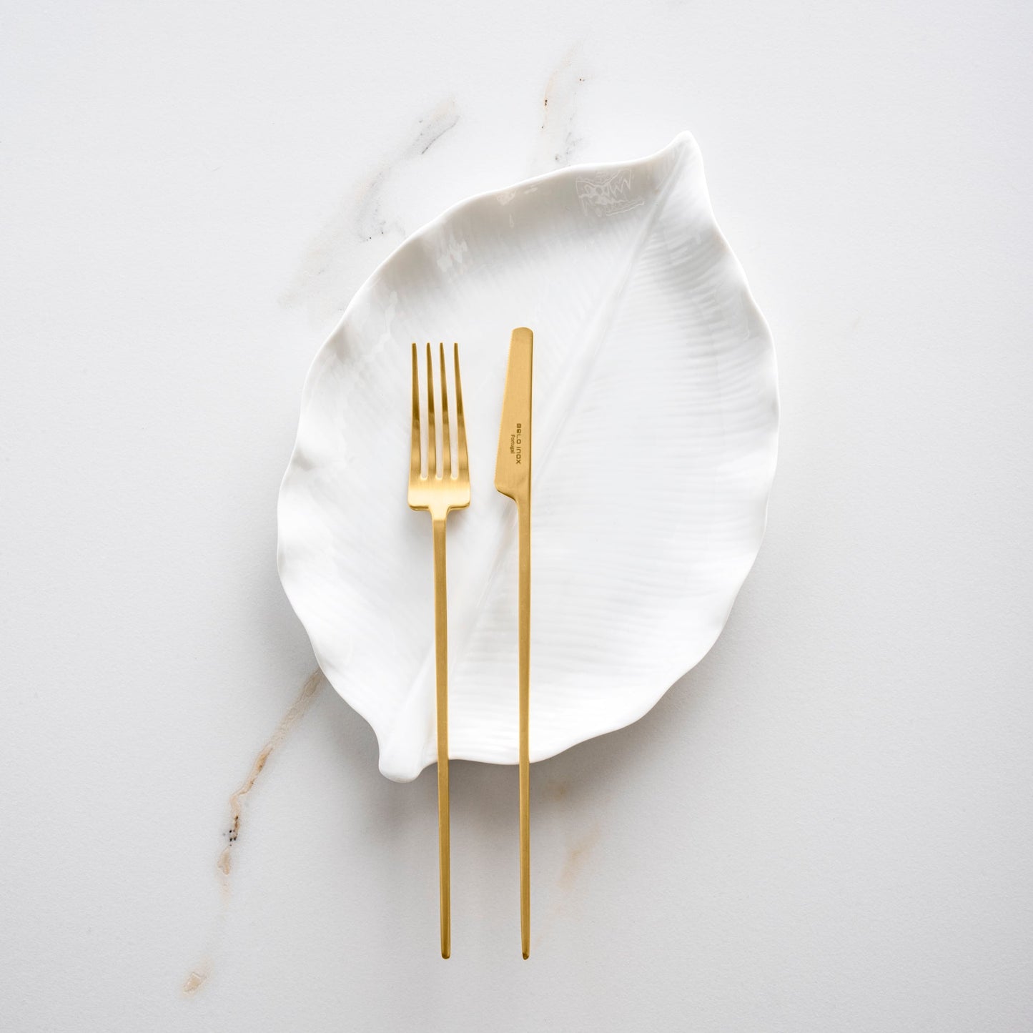 Belo Inox TWO gold matte cutlery set displayed on a white leaf-shaped plate over a marble surface.