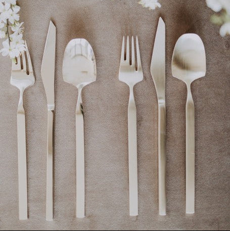 Belo Inox Vertice cutlery set in polished and brushed finishes, displayed on a soft neutral background with delicate white floral branches