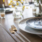 Cutipol Goa Grey Gold forks elegantly placed next to a decorative blue and white plate on a wooden dining table with floral arrangements.